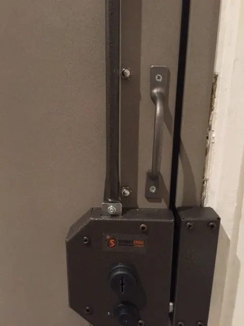 Serrure Securystar en applique posée sur une porte blindée, tringle et coffre visibles
